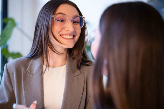 A woman in glasses smiles while talking to another person in a business setting.