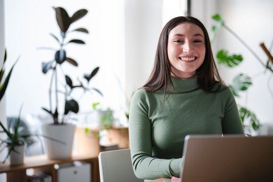 A young woman smiles while working on her laptop in a bright, plant-filled room.