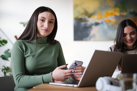 A young woman with dark hair and a nose ring looks at the camera while holding a smartphone. She is seated at a table with a laptop, and another person is working on a laptop in the background.