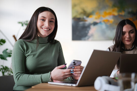 A young woman smiles while holding a smartphone and working on a laptop. Another person is visible in the background, also using a computer.
