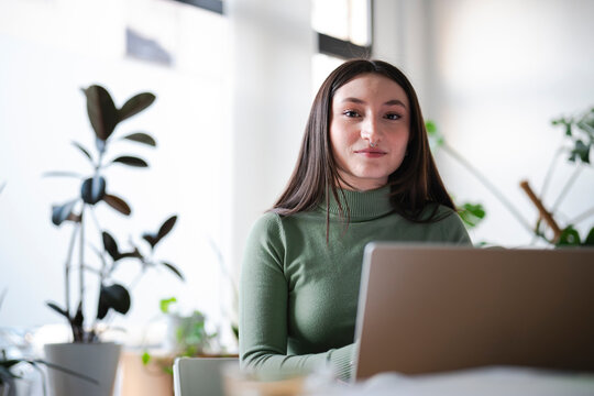 A young woman with dark hair and a nose ring sits at a desk with a laptop, looking directly at the camera. She is wearing a green turtleneck sweater.
