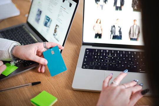 A person holds a debit card while shopping online on a laptop, with another laptop displaying clothing items in the background.