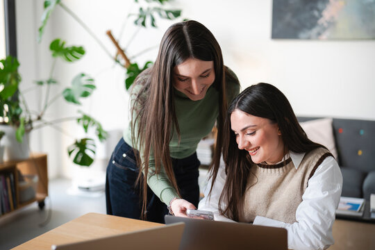 Two young women collaborate on a project, one showing the other something on a mobile device while working at a desk with laptops.
