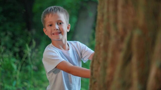 Boy emerges from behind tree playing hide and seek in forest. Child smiles during game near trunk outdoors. Kid plays hide and seek. Boy hides behind tree smiling. Child enjoys forest game.