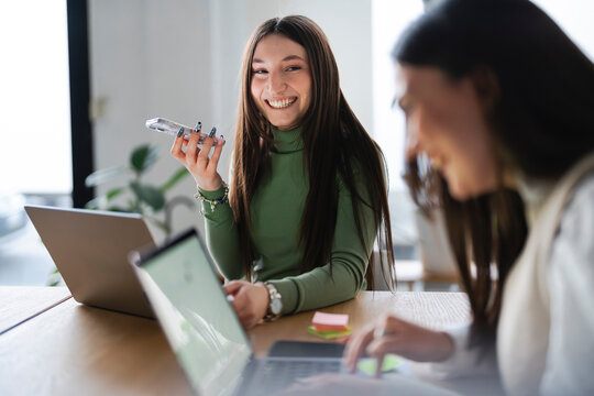 A young woman smiles while holding her phone, sitting at a table with laptops and sticky notes, suggesting a collaborative work or study session.