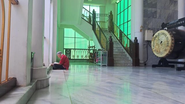 Close-up of a senior Asian Muslim man sitting in a corner of a mosque porch, opening and reading the Holy Quran. This serene footage captures the essence of aging with spiritual devotion.
