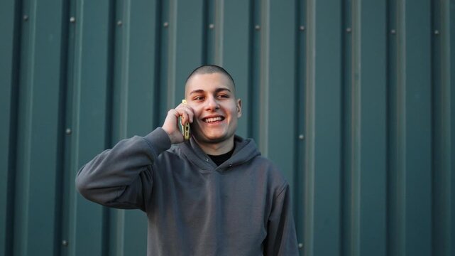Young smiling man with buzz cut wearing a hoodie talking on his smartphone against a corrugated metal wall outdoors