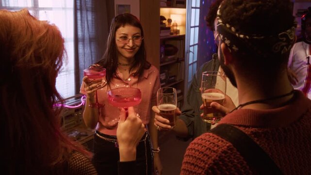 Over shoulder shot of smiling young adults toasting with drinks while casually socializing at home party enjoying conversation and DJ music set in neon lit living room