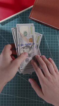 Vertical close-up of hands quickly counting a stack of one-hundred dollar bills totaling 1400 USD on a professional workspace.