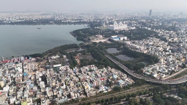 Expansive aerial view of Hyderabad featuring Hussain Sagar, dense urban neighborhoods, and iconic landmarks under a hazy daytime sky. The Telugu Thalli Flyover and NTR Marg roads are used by vehicles
