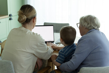 Obraz premium Three generations sitting together at home using laptop. Young caregiver helping senior woman and child with digital technology. Concept of family support, intergenerational learning and modern lifest