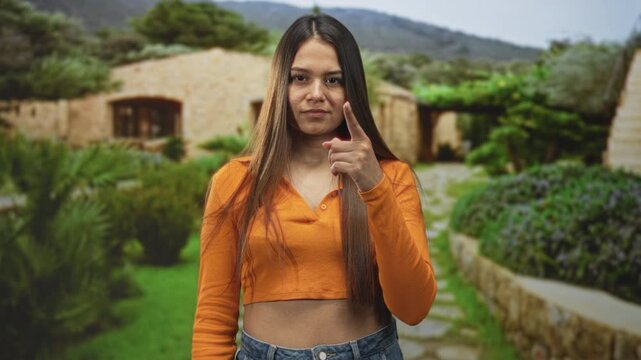 Young brunette woman points finger at camera in front of stone building, wearing orange cropped top and jeans, assertive stare; confidence challenge.
