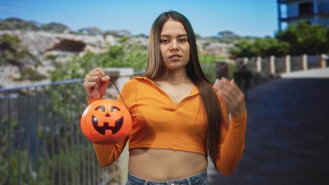 Woman holds a jack o lantern pumpkin bucket by hand on a coastal promenade street with railing, stone cliffs and distant building; festive.