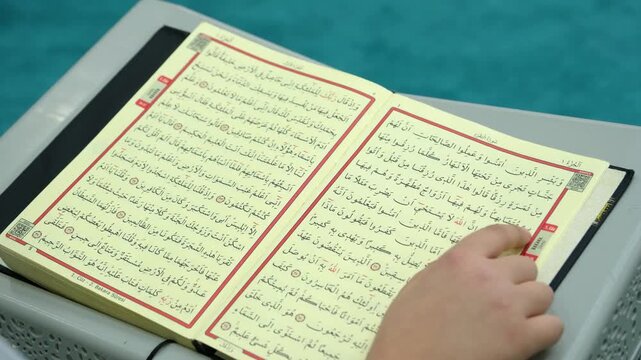 Close up of the holy book Quran on a wooden stand with hands moving across the Arabic text for religious education and spiritual study concepts indoors at a mosque or school