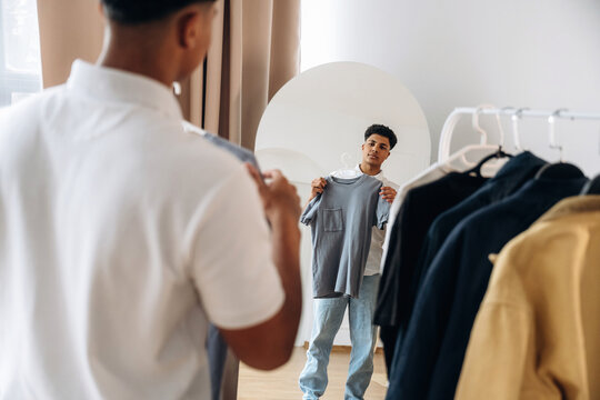 Young man holding gray t-shirt while looking at himself in a mirror, surrounded by various clothing items hanging on a rack in a bright, modern room
