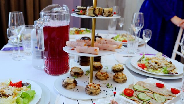 Plates filled with salads, snacks, ham, and stuffed mushrooms arranged on a three tiered stand with wine glasses and a juice pitcher, creating an elegant catering setup