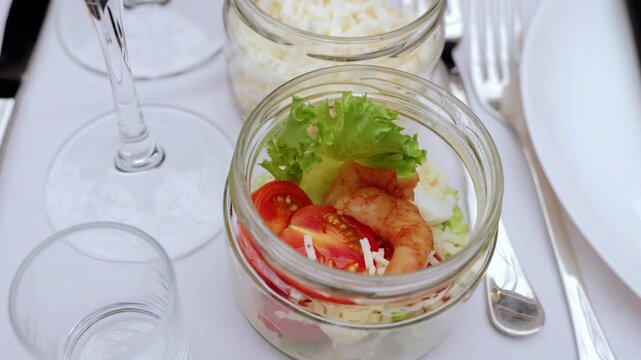 Glass jars displaying layered caesar salad featuring grilled shrimp, vibrant cherry tomatoes, crisp lettuce, and grated parmesan nestled on pristine white tablecloth