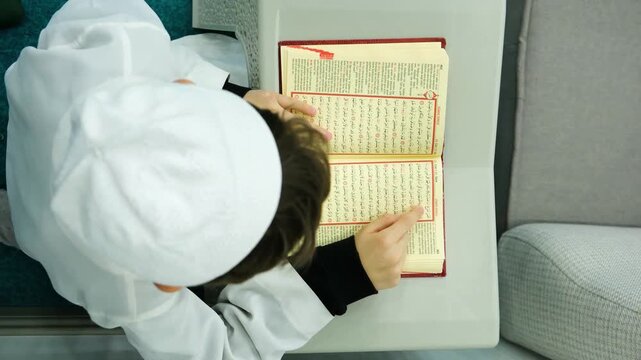 Overhead view of a young boy wearing a white prayer cap reading the holy Quran while sitting on a teal carpet for religious education and islamic school concept at daytime