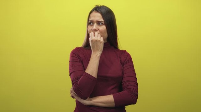 Woman biting nails with hand to mouth in studio, maroon turtleneck, folded arm and yellow backdrop visible; anxiety coping.