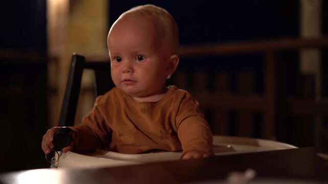 Mother picks up a dropped water bottle while her baby plays with keys in a high chair at an evening restaurant.