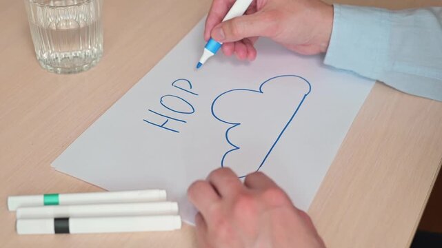 A person writes the word Hope using colorful markers on a sheet of paper. The activity occurs on a wooden table, with glasses of water and markers present