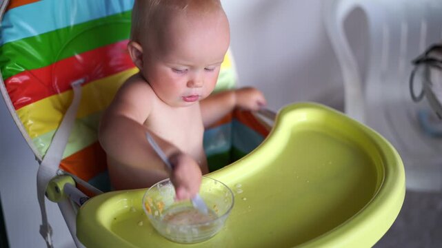 Curious messy toddler sitting in a high chair learns to eat cereal with a spoon causing happy mealtime chaos.