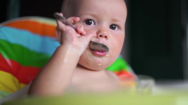 Close up of a joyful messy baby eating cereal with hands and a spoon during a chaotic fun breakfast.