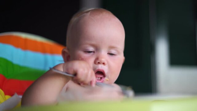 Cute messy infant independently explores eating cereal from a bowl developing fine motor skills in a high chair.