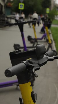 Close-up view of electric scooters lined along a city sidewalk, handlebars in sharp focus with shallow depth of field, while pedestrians and street signs blur in the background