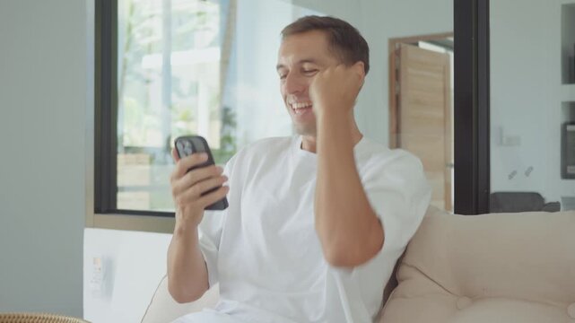 Success man celebrating with smartphone reading good news message and smiling at home on couch. Success concept shows emotional reaction to positive message and mobile success communication