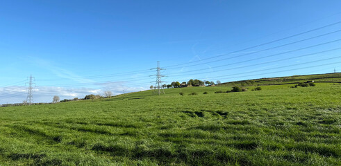 Obraz premium A lush green field stretches under a clear blue sky. Tall pylons tower over the landscape, connecting distant hills near the old village of Thornton, Yorkshire, UK