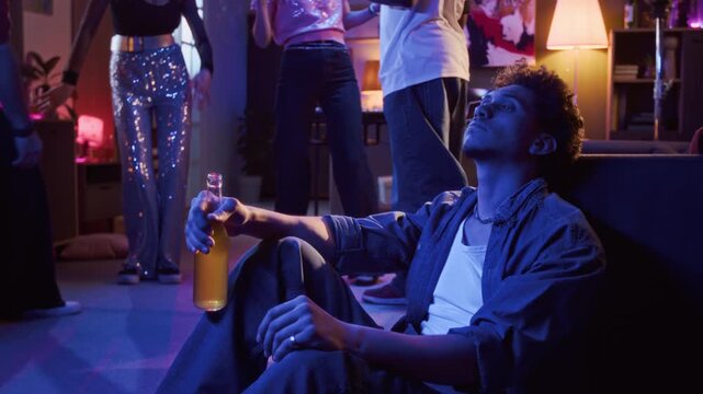 Multiethnic young adult man sitting on floor with bottle of drink in hand feeling overwhelmed during home party while friends having fun on dance floor