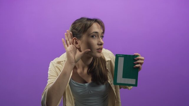 Woman holding green learner l plate and hand to ear listening gesture in purple studio setting; learning practice attentive.