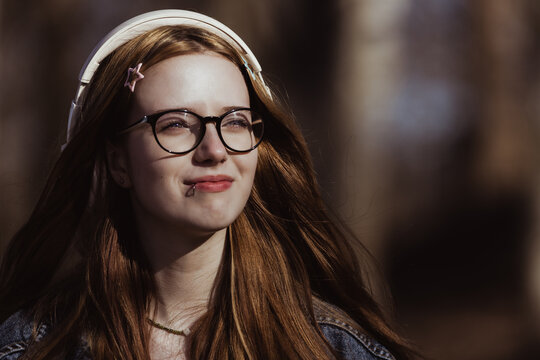Young woman with long red hair wearing glasses and headphones stands outdoors in a natural setting, sunlight filtering through trees in the background