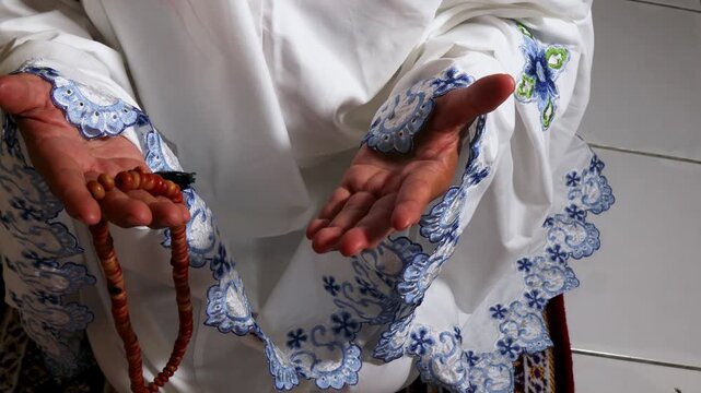 a Muslim woman is praying using a mukena and prayer beads