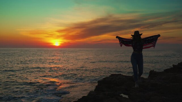 Proud american woman wear cowboy hat stand on rocky cliff, hold united states flag high against dramatic sunset over ocean, symbolizing patriotism and freedom.Usa celebrate 4th july. Independence day