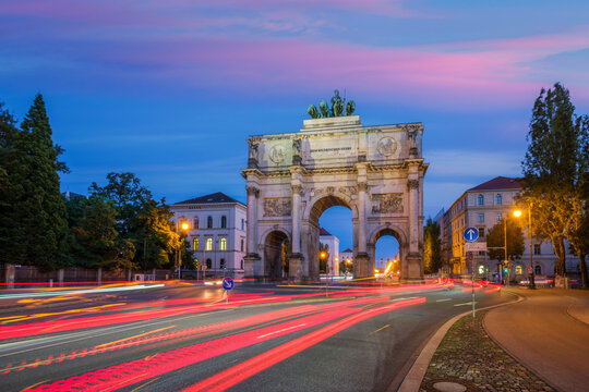 Long exposure of the Siegestor (Victory Gate) in Munich, Germany, at twilight with colorful car light trails and sunset sky.
