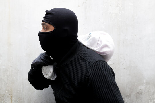 Man in black balaclava holds white sack. He looks directly at camera with intense gaze. His gloved hand grips the sack tightly. Urban concrete wall forms neutral background. Image conveys tension
