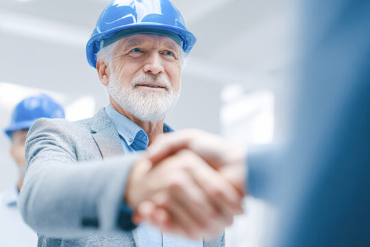 Senior man in a blue hard hat shaking hands, symbolizing agreement or partnership at a construction site.
