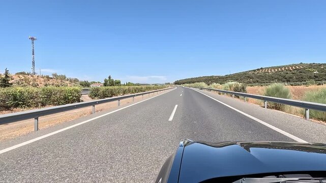 A car driving along a straight rural highway under a clear blue sky, capturing open road scenery, guardrails, hills, and peaceful countryside view