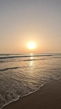 Vertical hand-shot video of the beautiful sunrise or sunset over the ocean horizon, with a clear sky during the golden hour and gentle waves breaking on the sandy shore of the beach, creating a calm a