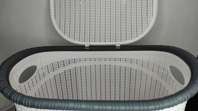 A close-up shot of colorful clothes being thrown into an empty white laundry basket with a woven texture, followed by a hand closing the lid against a grey wall background.