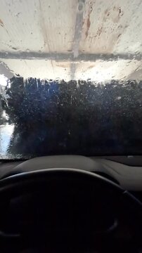 Vertical video of POV from inside a car during automatic wash tunnel, blue foam and water covering the windshield, dashboard visible, brushes and light outside