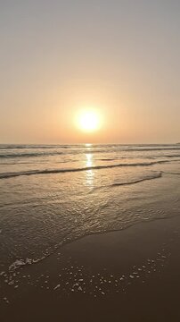 Vertical video of a person walking on the beach sand towards sunset or sunrise over the ocean horizon, with a clear sky during the golden hour. First-person view