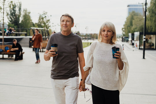 Adult mature happy romantic couple in love outdoors in city street park. A blonde caucasian man and woman spend time together, drinking coffee. Senior wife and husband walking outside on town square