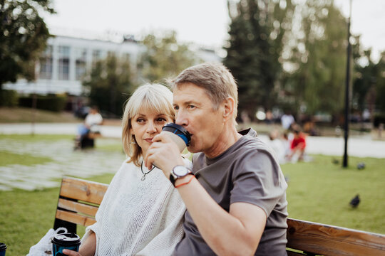 An adult mature happy couple in love sitting on bench outdoors in city street park. A blonde caucasian man and woman spend time together and drinking coffee. Senior wife and husband walking outside