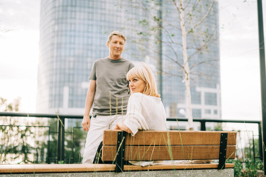 An adult mature happy romantic couple in love sitting on bench outdoors in city street park. A blonde caucasian man and woman spend time together. Senior wife and husband walking outside