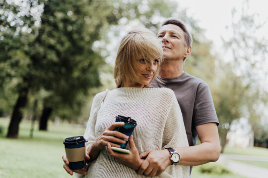 An adult mature happy couple in love hugging outdoors in city park. A blonde caucasian man and woman spend time together and drinking coffee. Senior wife and husband walking outside