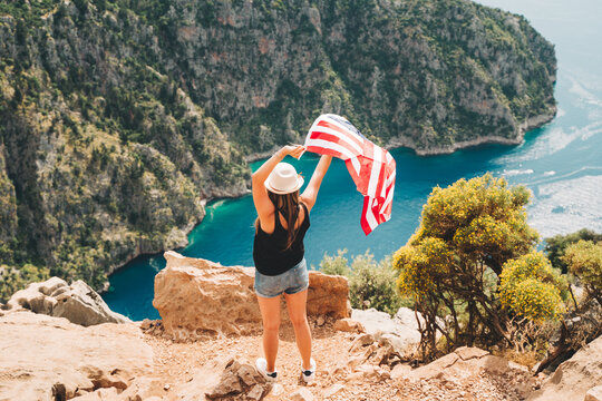 Young woman standing on a rock cliff and waving the US flag while looking at sea beneath. Girl traveller waving the American flag while standing on a mountain top. 4 fourth July Independence Day