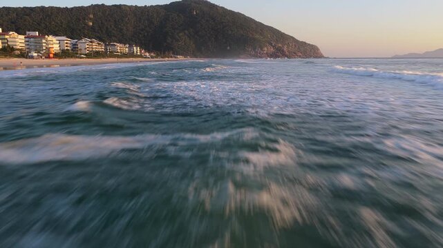 Low-altitude drone skimming crashing waves toward Praia Brava shore, luxury homes and headland at golden hour.